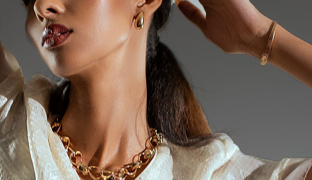 A model’s profile from the lips down, with a gold drop earring, gold bangle and gold chain.
