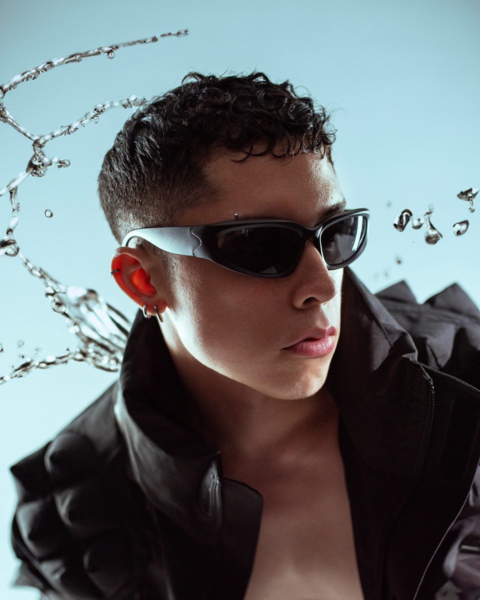 A model in wraparound black sunglasses and an oversized black jacket, with water splashing against a pale blue backdrop.