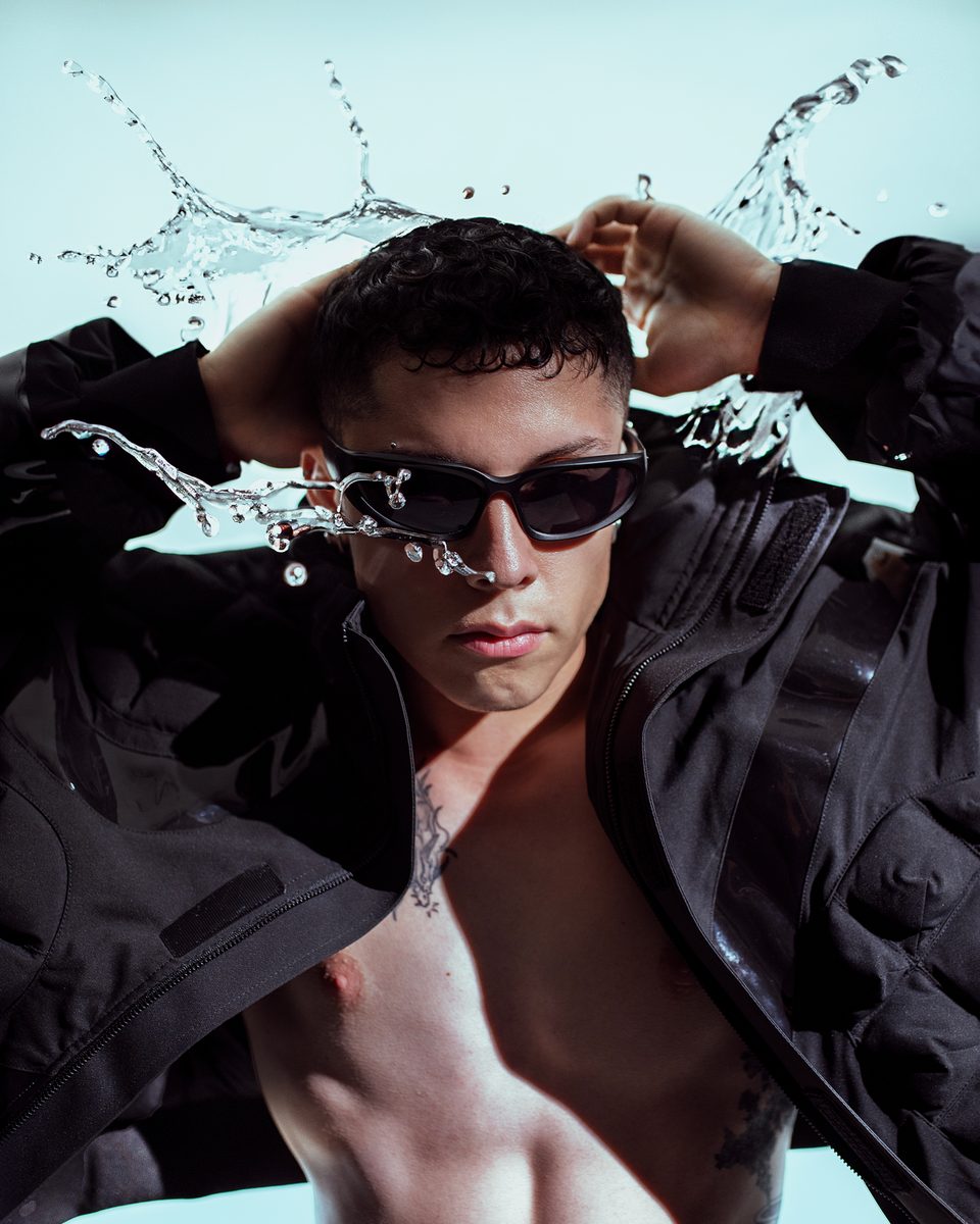 A model in a black quilted jacket and sunglasses, arms raised as water splashes behind him.