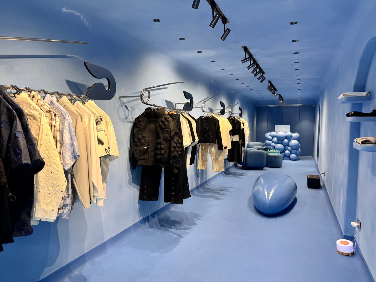 Clothes on oversized safety-pin rails along a blue wall, with a smooth blue form on the floor.